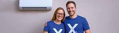 Zwei Personen mit einem X auf blauen Shirts stehen vor einem Klimagerät